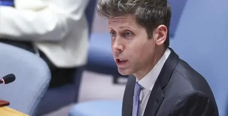 Hombre con traje oscuro y corbata azul hablando con expresión seria, sentado en una sala de conferencias con sillas azules al fondo.
