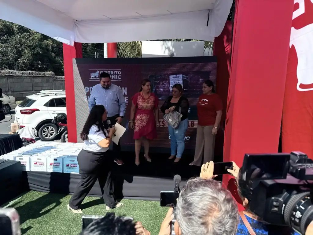 Presentadora de Distrito Carnic con micrófono anuncia a los ganadores frente a cajas de premios y cámaras de prensa