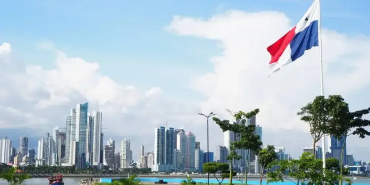 Vista panorámica de un moderno skyline de rascacielos junto a la costa, con una bandera nacional ondeando en primer plano y un barco en el agua.