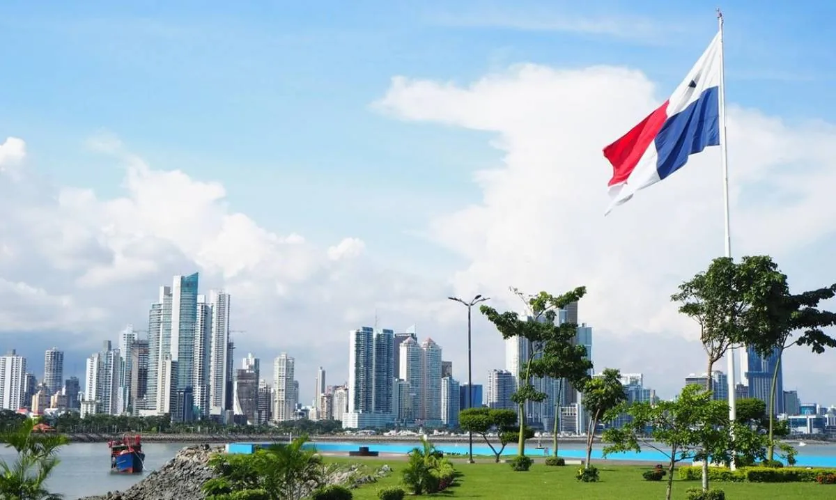 Vista panorámica de un moderno skyline de rascacielos junto a la costa, con una bandera nacional ondeando en primer plano y un barco en el agua.