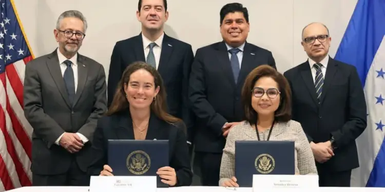 Dos mujeres sentadas firmando documentos oficiales acompañadas por cuatro hombres en traje y banderas.