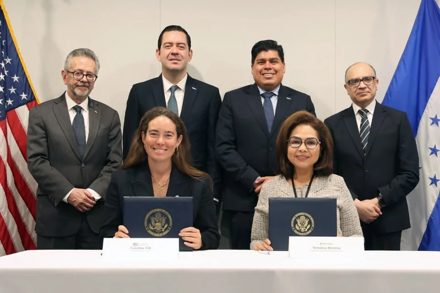 Dos mujeres sentadas firmando documentos oficiales acompañadas por cuatro hombres en traje y banderas.