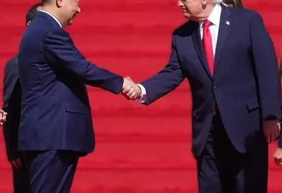 Dos hombres de negocios, uno con traje azul oscuro y corbata roja, el otro con traje azul, se dan la mano sobre una alfombra roja, simbolizando un acuerdo o negociación.