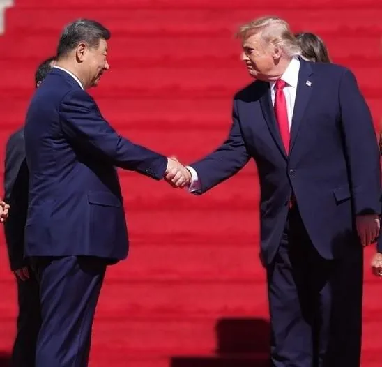Dos hombres de negocios, uno con traje azul oscuro y corbata roja, el otro con traje azul, se dan la mano sobre una alfombra roja, simbolizando un acuerdo o negociación.