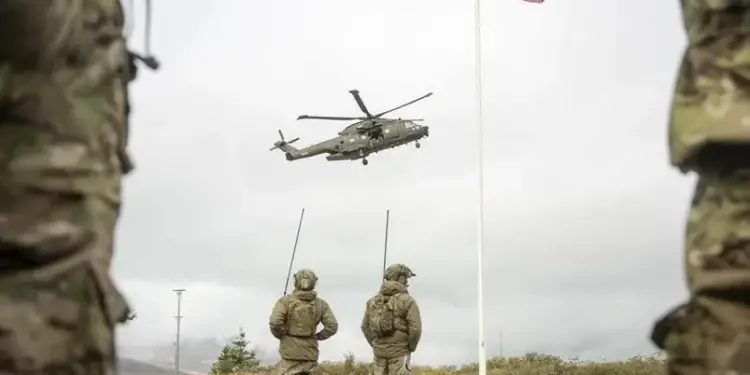 Helicóptero militar verde oscuro en vuelo sobre dos personas con uniformes de camuflaje y equipos de comunicación, observando. Una bandera roja y blanca ondea en un mástil.