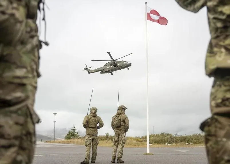 Helicóptero militar verde oscuro en vuelo sobre dos personas con uniformes de camuflaje y equipos de comunicación, observando. Una bandera roja y blanca ondea en un mástil.
