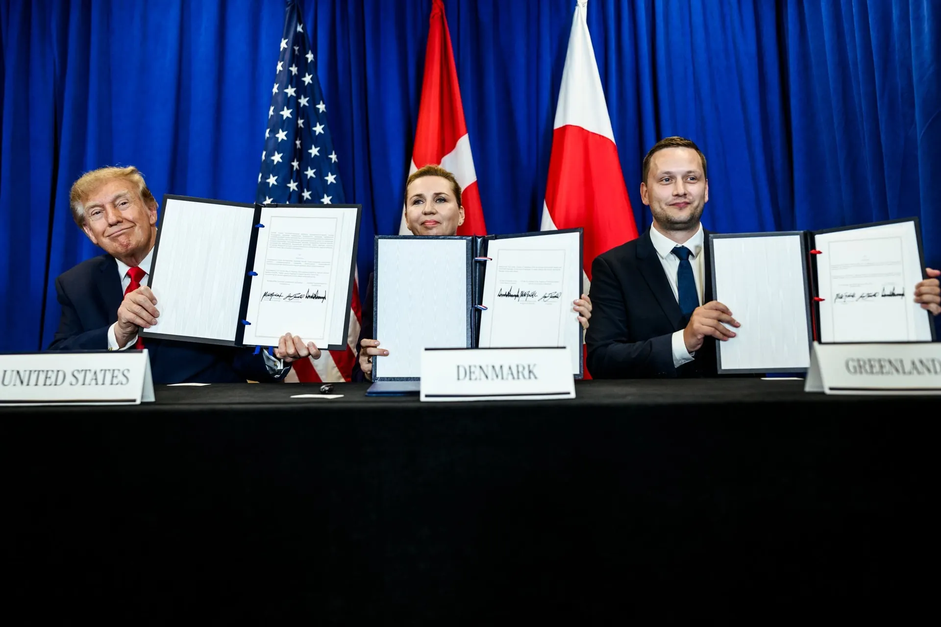 Tres personas sentadas ante una mesa con carteles de United States, Denmark y Greenland mostrando documentos firmados.
