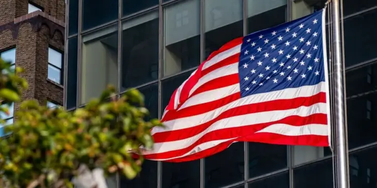 Bandera de los Estados Unidos ondeando en su asta frente a la fachada de un edificio moderno de cristal.