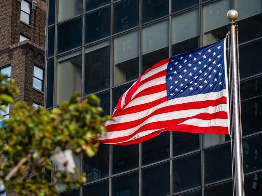 Bandera de los Estados Unidos ondeando en su asta frente a la fachada de un edificio moderno de cristal.