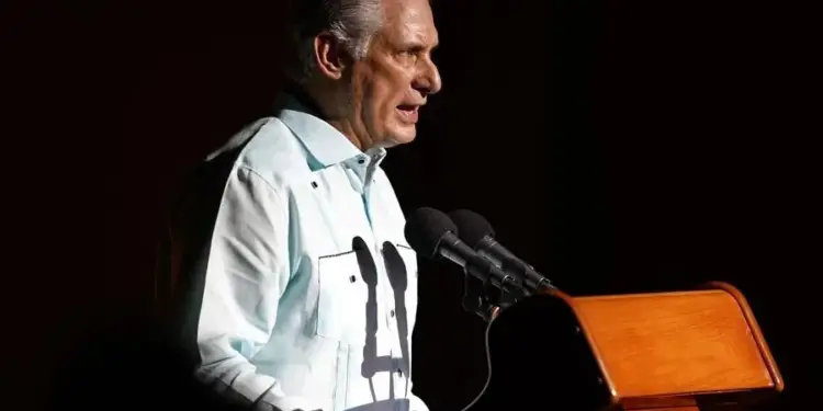 Hombre de cabello gris en guayabera clara, hablando en un podio de madera con dos micrófonos. Parece dar un discurso o presentación.