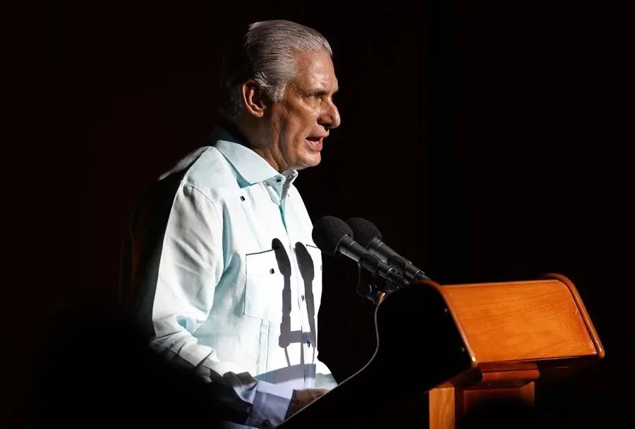 Hombre de cabello gris en guayabera clara, hablando en un podio de madera con dos micrófonos. Parece dar un discurso o presentación.