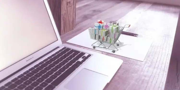 Laptop plateada con pantalla en blanco, un carrito de compras miniatura lleno de regalos coloridos, un papel y un bolígrafo sobre una mesa de madera. Representa compras en línea.