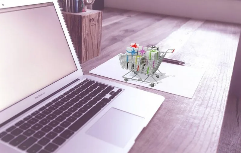 Laptop plateada con pantalla en blanco, un carrito de compras miniatura lleno de regalos coloridos, un papel y un bolígrafo sobre una mesa de madera. Representa compras en línea.