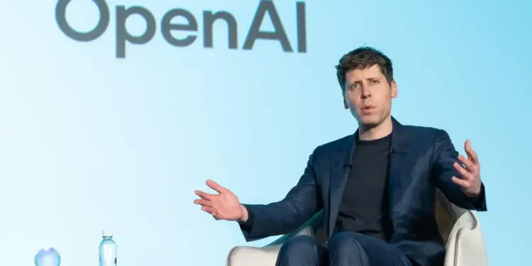 Hombre con traje azul oscuro hablando con las manos extendidas, con el logo de OpenAI al fondo.