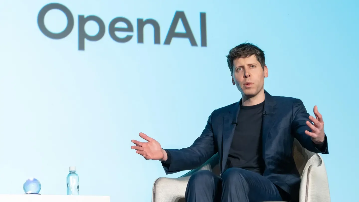 Hombre con traje azul oscuro hablando con las manos extendidas, con el logo de OpenAI al fondo.