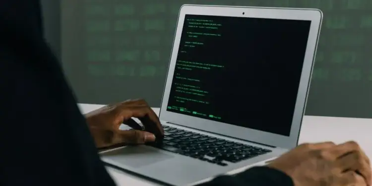 Persona con traje oscuro escribiendo código en un portátil con pantalla verde brillante, con fondo de datos digitales en verde.