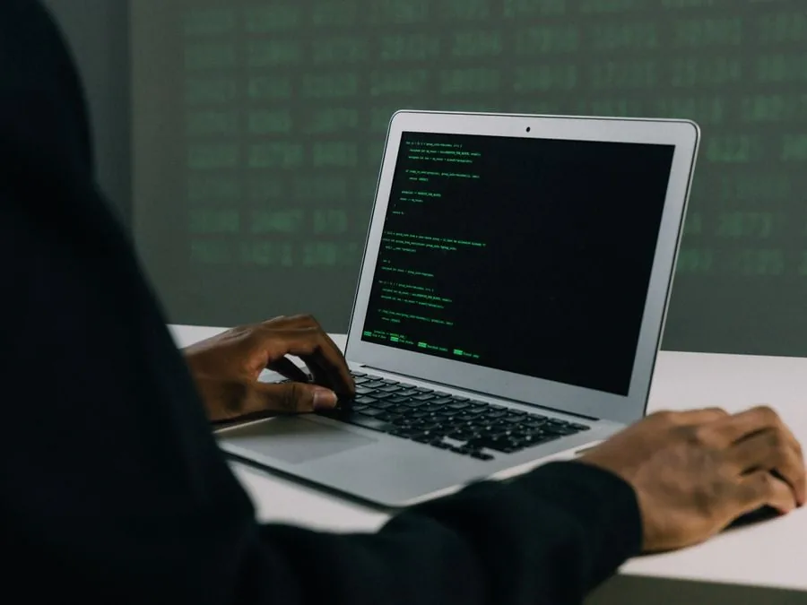 Persona con traje oscuro escribiendo código en un portátil con pantalla verde brillante, con fondo de datos digitales en verde.