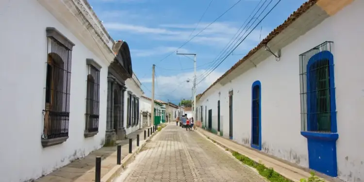 Vista de una calle empedrada bordeada de casas blancas coloniales con ventanas enrejadas bajo un cielo azul con nubes.