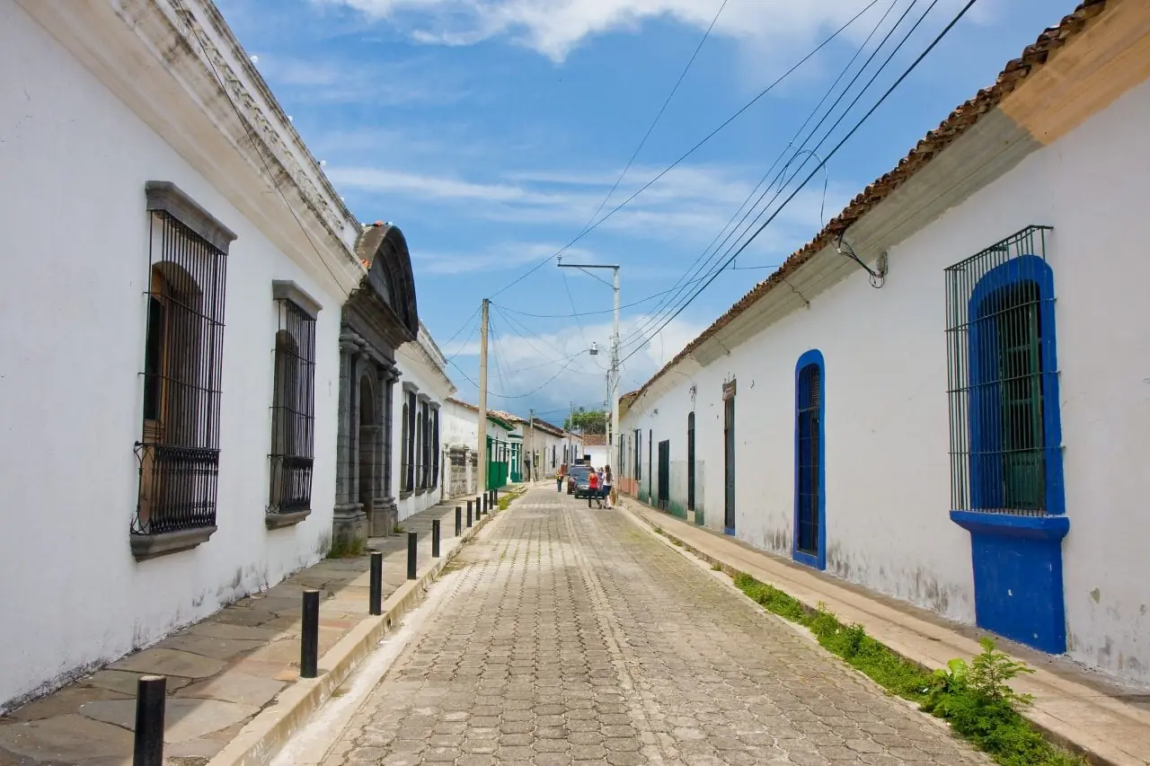 Vista de una calle empedrada bordeada de casas blancas coloniales con ventanas enrejadas bajo un cielo azul con nubes.