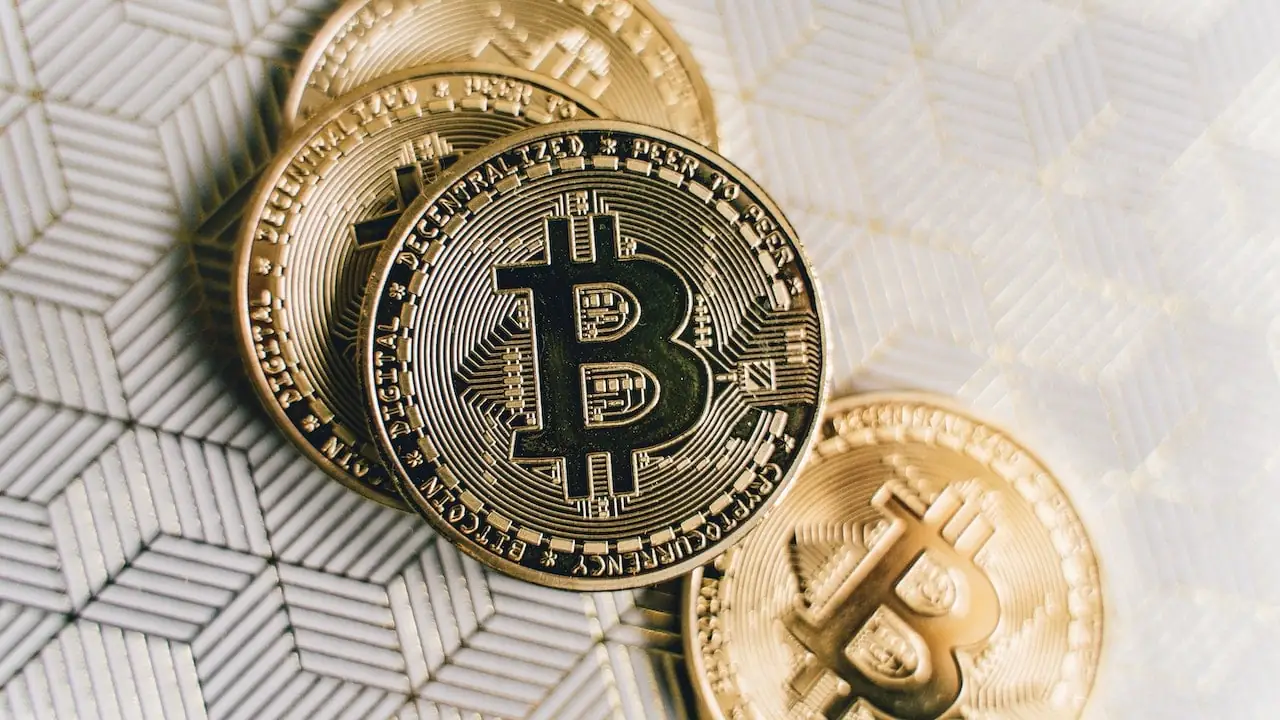 Monedas físicas doradas de bitcoin dispuestas sobre una superficie con patrón geométrico texturizado en tonos claros.