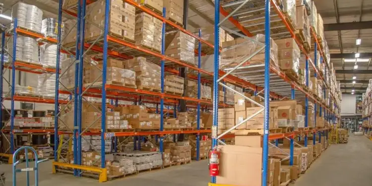 Amplio almacén logístico con altas estanterías metálicas azules y naranjas llenas de cajas y mercancía organizada.