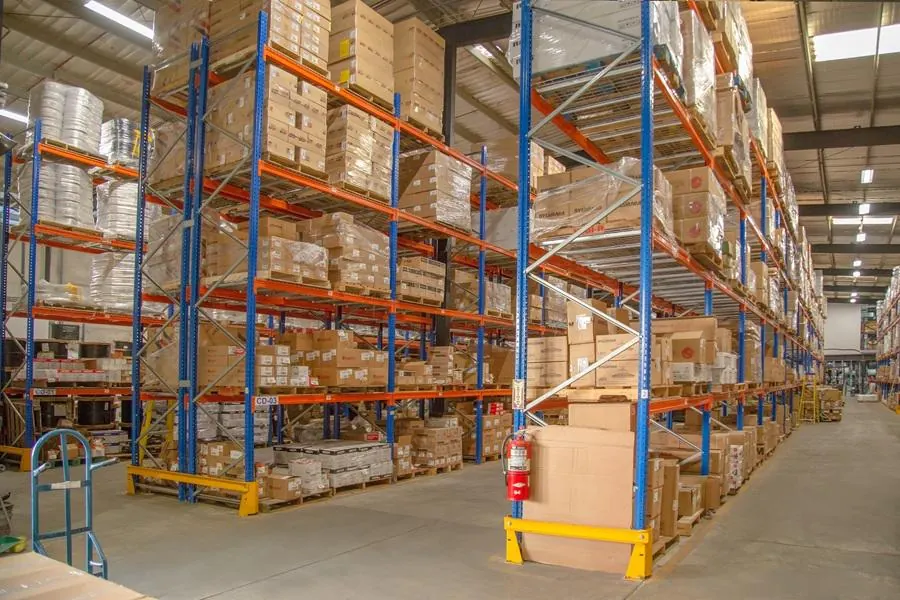 Amplio almacén logístico con altas estanterías metálicas azules y naranjas llenas de cajas y mercancía organizada.