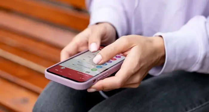 Primer plano de manos sosteniendo un smartphone con funda lila, interactuando con una aplicación de correo o mensajería. La persona viste camisa lila y jeans oscuros, sentada en un banco de madera.