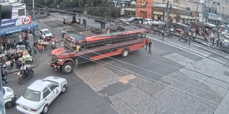 Un autobús rojo y negro en un cruce de calles urbanas junto a comercios locales, vehículos y peatones.