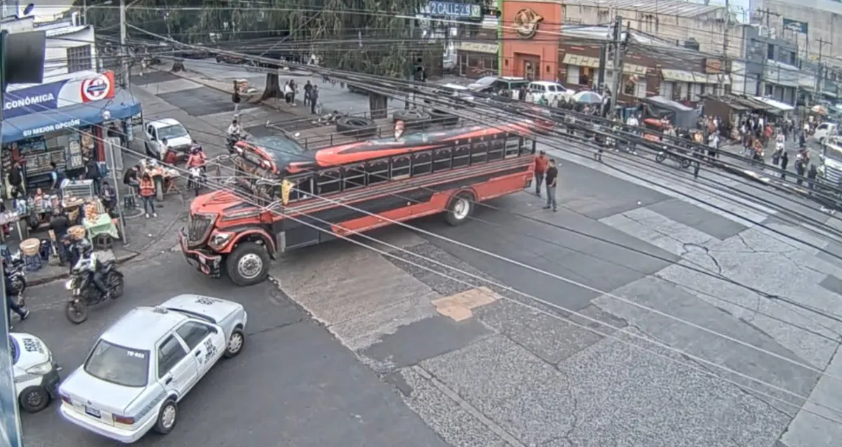Un autobús rojo y negro en un cruce de calles urbanas junto a comercios locales, vehículos y peatones.