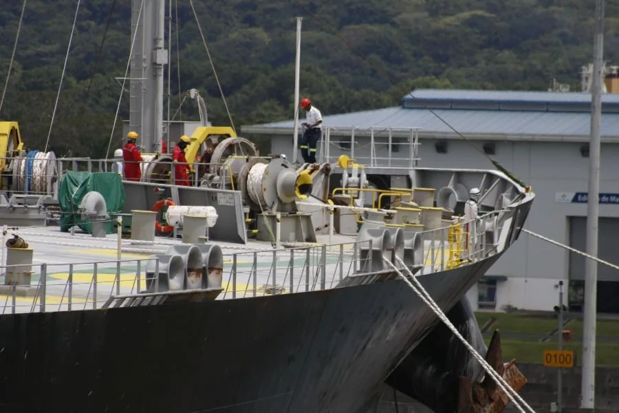 Vista de la cubierta de un gran barco con trabajadores en puerto y equipamiento industrial.