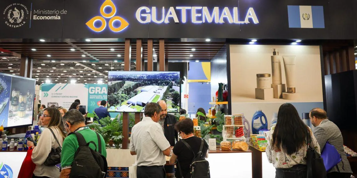 Stand de Guatemala en una feria de negocios con el logo del Ministerio de Economía. Personas interactúan y observan productos y pantallas con imágenes de instalaciones industriales.