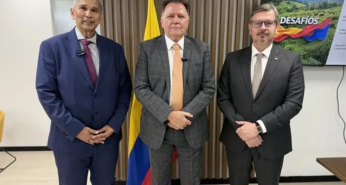 Tres hombres con traje formal de pie en una oficina frente a una bandera.