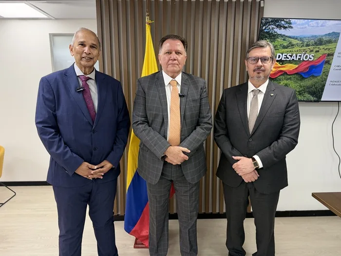 Tres hombres con traje formal de pie en una oficina frente a una bandera.