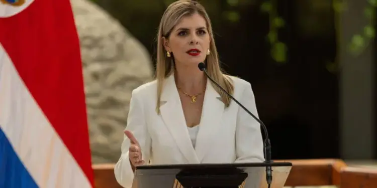 Mujer profesional en blazer blanco dando un discurso en podio con micrófono. Bandera de Costa Rica visible. Representa liderazgo económico.