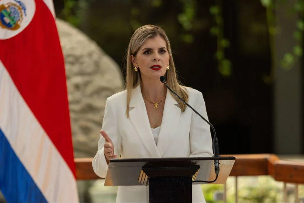 Mujer profesional en blazer blanco dando un discurso en podio con micrófono. Bandera de Costa Rica visible. Representa liderazgo económico.
