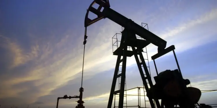 Silueta de una bomba de extracción de petróleo (pumpjack) contra un cielo azul y anaranjado al amanecer o atardecer, con tuberías rojas en primer plano.