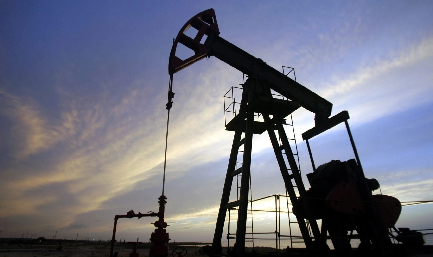 Silueta de una bomba de extracción de petróleo (pumpjack) contra un cielo azul y anaranjado al amanecer o atardecer, con tuberías rojas en primer plano.