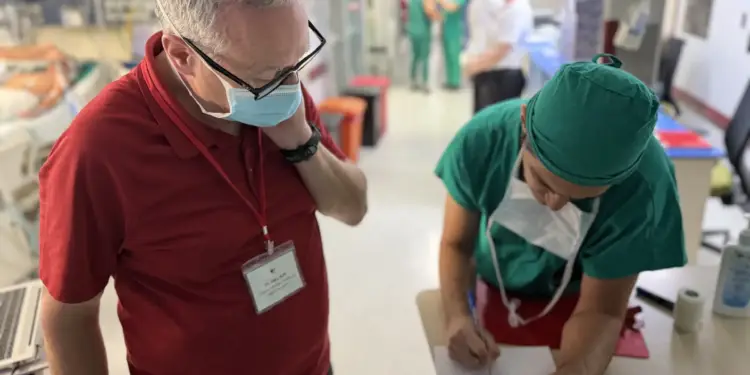 Dos profesionales sanitarios con mascarilla en el pasillo de un hospital, uno escribe y otro observa.