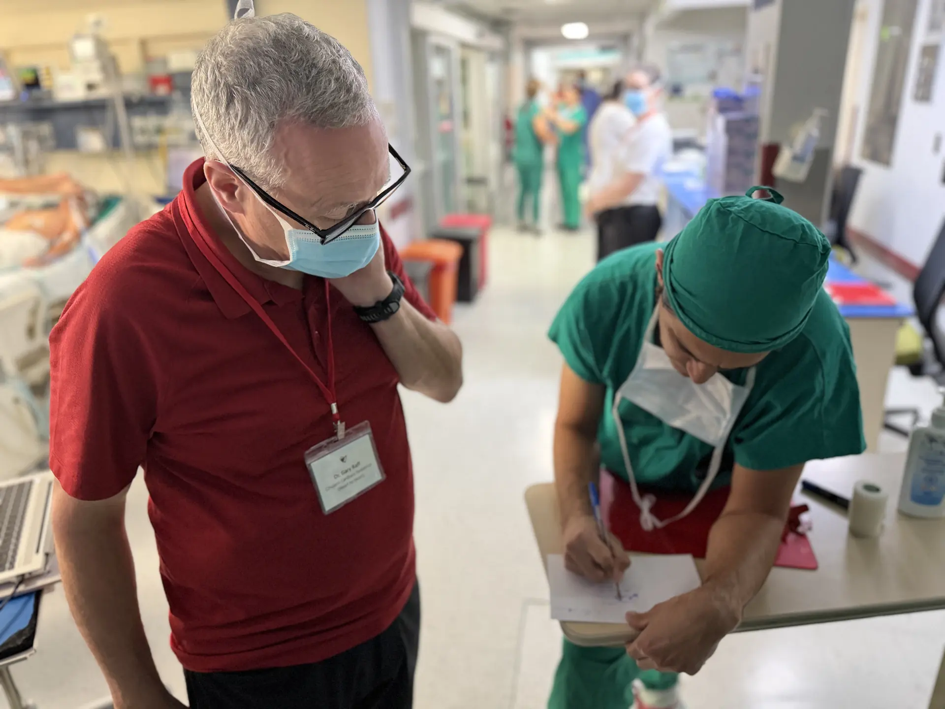 Dos profesionales sanitarios con mascarilla en el pasillo de un hospital, uno escribe y otro observa.