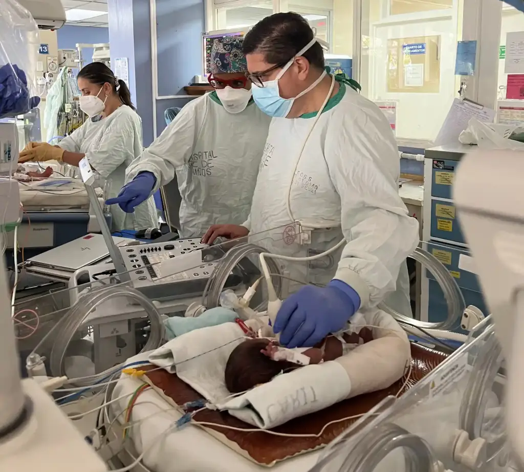 Personal médico con mascarillas y batas revisando a un bebé en una incubadora hospitalaria con equipos médicos avanzados.