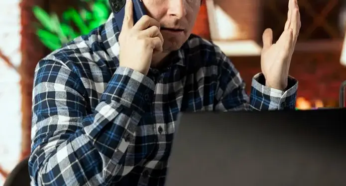 Hombre caucásico en camisa a cuadros azul y blanco hablando por teléfono móvil con expresión preocupada, gesticulando. Un portátil negro y auriculares azules están sobre el escritorio. Ambiente de oficina o trabajo remoto.