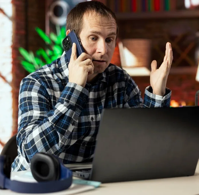 Hombre caucásico en camisa a cuadros azul y blanco hablando por teléfono móvil con expresión preocupada, gesticulando. Un portátil negro y auriculares azules están sobre el escritorio. Ambiente de oficina o trabajo remoto.