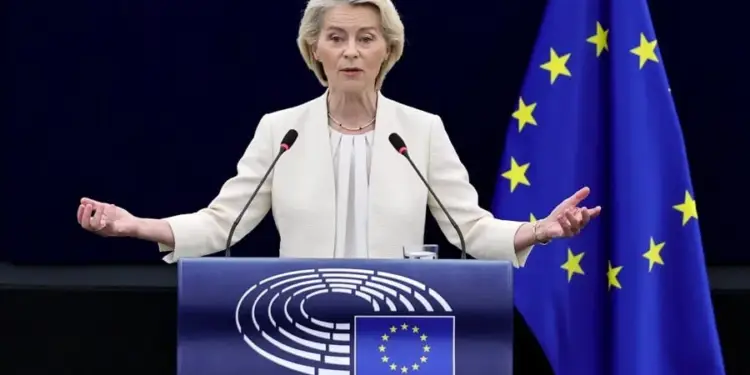 Mujer rubia con traje claro dando un discurso en un podio azul con el logo del Parlamento Europeo y la bandera de la UE. Gesticula con las manos abiertas, con dos micrófonos y un vaso de agua.