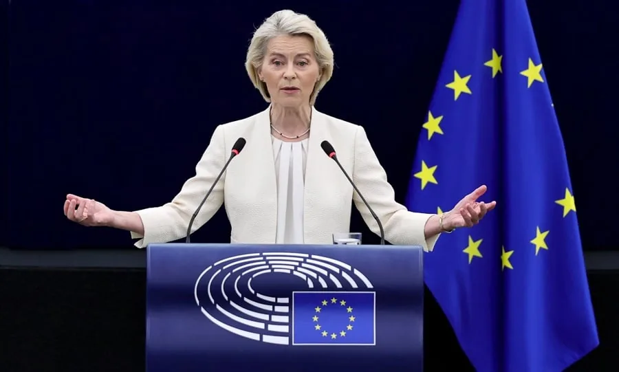 Mujer rubia con traje claro dando un discurso en un podio azul con el logo del Parlamento Europeo y la bandera de la UE. Gesticula con las manos abiertas, con dos micrófonos y un vaso de agua.
