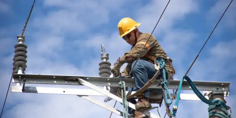 Trabajador con casco amarillo realizando mantenimiento en una estructura de alta tensión contra el cielo azul.