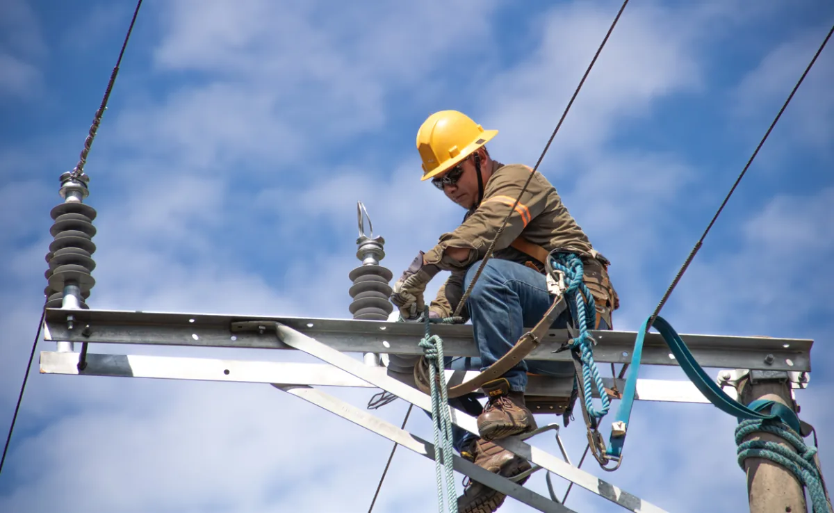 Trabajador con casco amarillo realizando mantenimiento en una estructura de alta tensión contra el cielo azul.