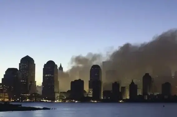 Vista de una ciudad con rascacielos al anochecer, donde una gran columna de humo oscuro se eleva sobre los edificios, reflejándose en el agua.