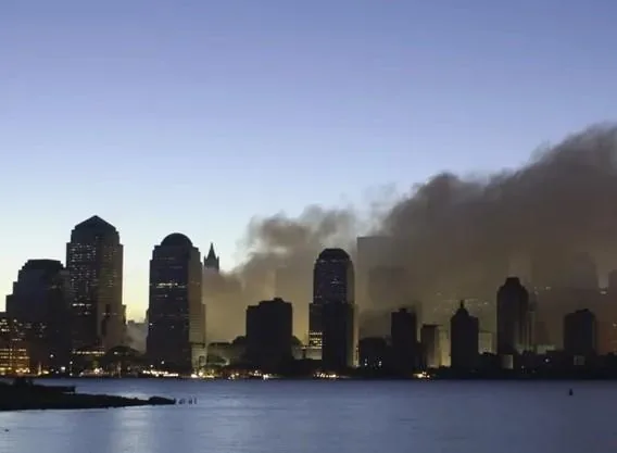 Vista de una ciudad con rascacielos al anochecer, donde una gran columna de humo oscuro se eleva sobre los edificios, reflejándose en el agua.