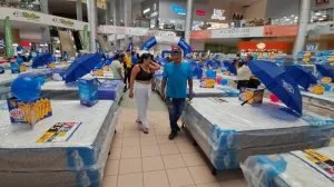 Personas caminando entre colchones en exhibición dentro de un centro comercial con decoraciones azules.
