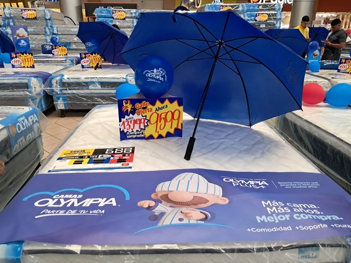 Exhibición de colchones en una tienda con paraguas azul abierto, globos y letreros de precios y ofertas especiales.